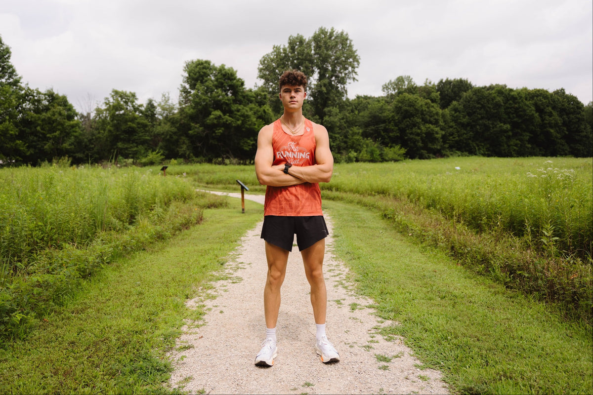 Dominic (The Running Effect) stands on a gravel path in a grassy field wearing an orange singlet with 'The Running Effect' printed on the front, black athletic shorts, and white sneakers.