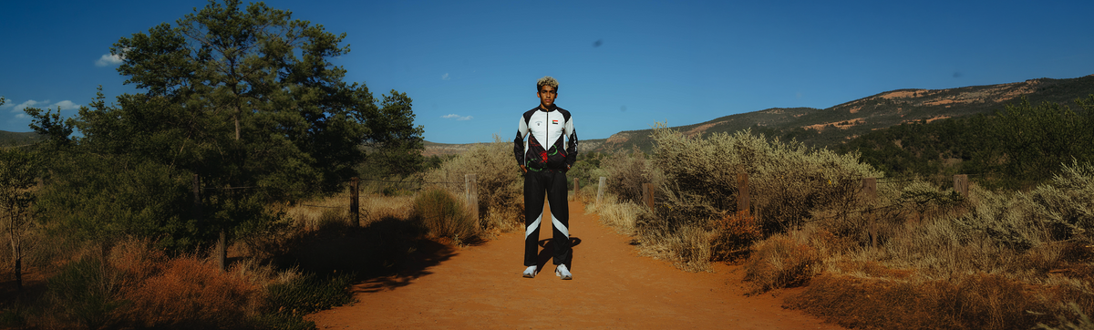 Yaseen Abdalla stands centered on a wide dirt path in an outdoor desert landscape with shrubs, trees, and hills under a clear blue sky.